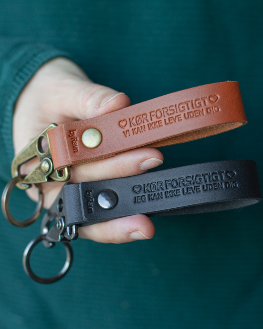 Zwei HOOK Schlüsselanhänger aus Cognac- und schwarzem Leder in der Hand gehalten mit der Gravur "Kør forsigtigt ♡ vi kan ikke leve uden dig"
