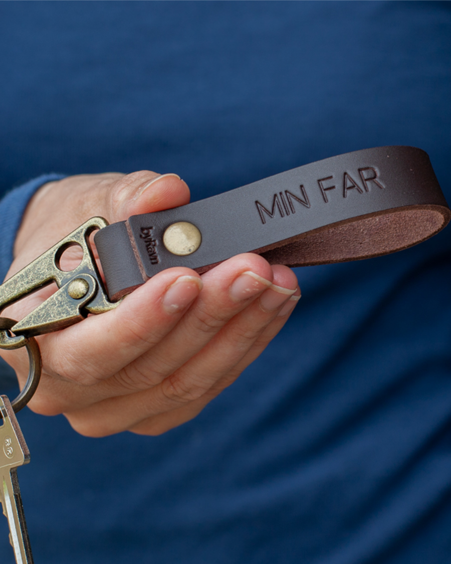 HOOK Schlüsselanhänger aus dunkelbraunem Leder mit Karabiner in Antikmessing und der Gravur "Min far", in der Hand gehalten mit Schlüssel