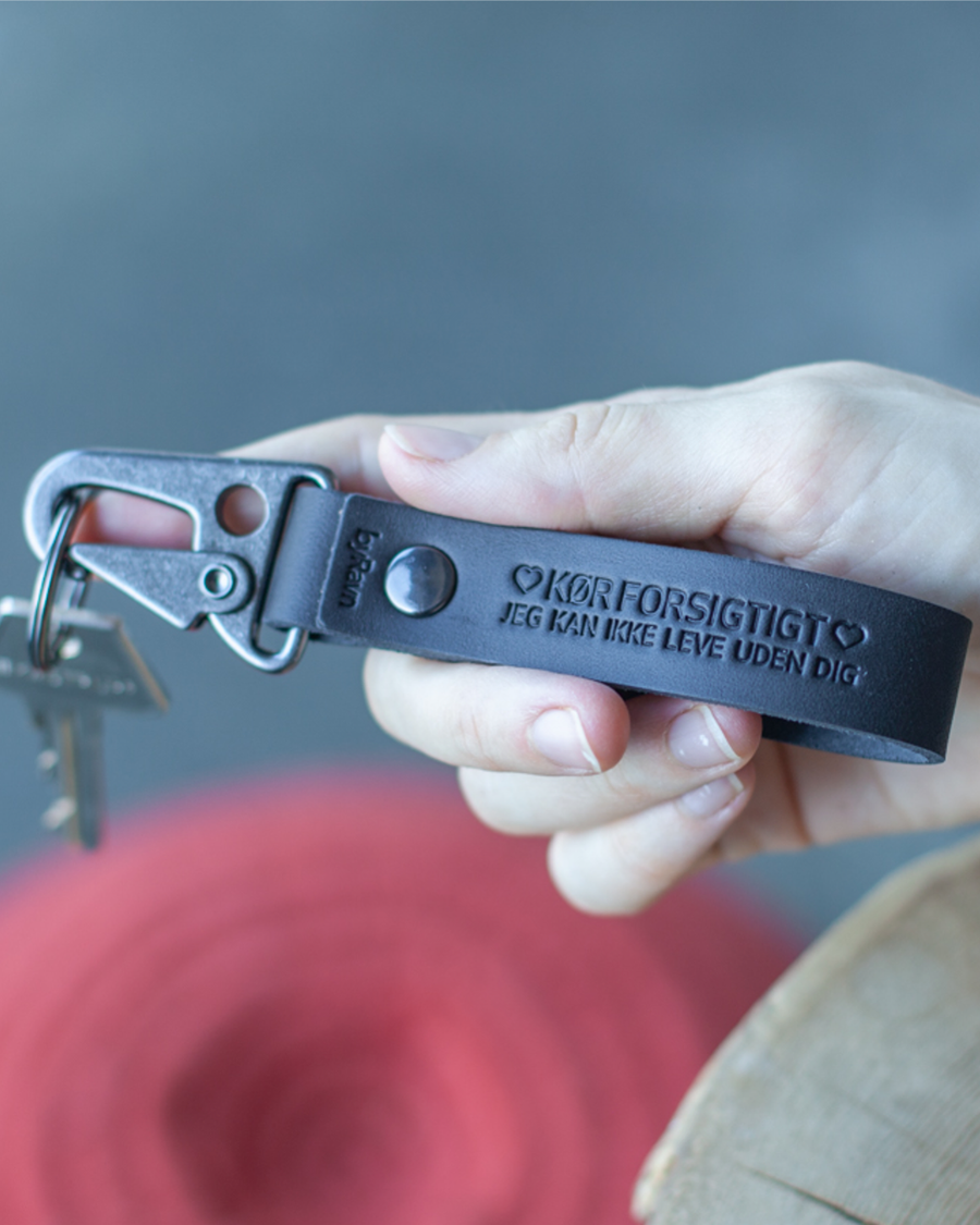 HOOK Schlüsselanhänger aus schwarzem Leder mit schwarzem Karabiner und der Gravur "Kør forsigtigt – jeg kan ikke leve uden dig" ♡ in der Hand
