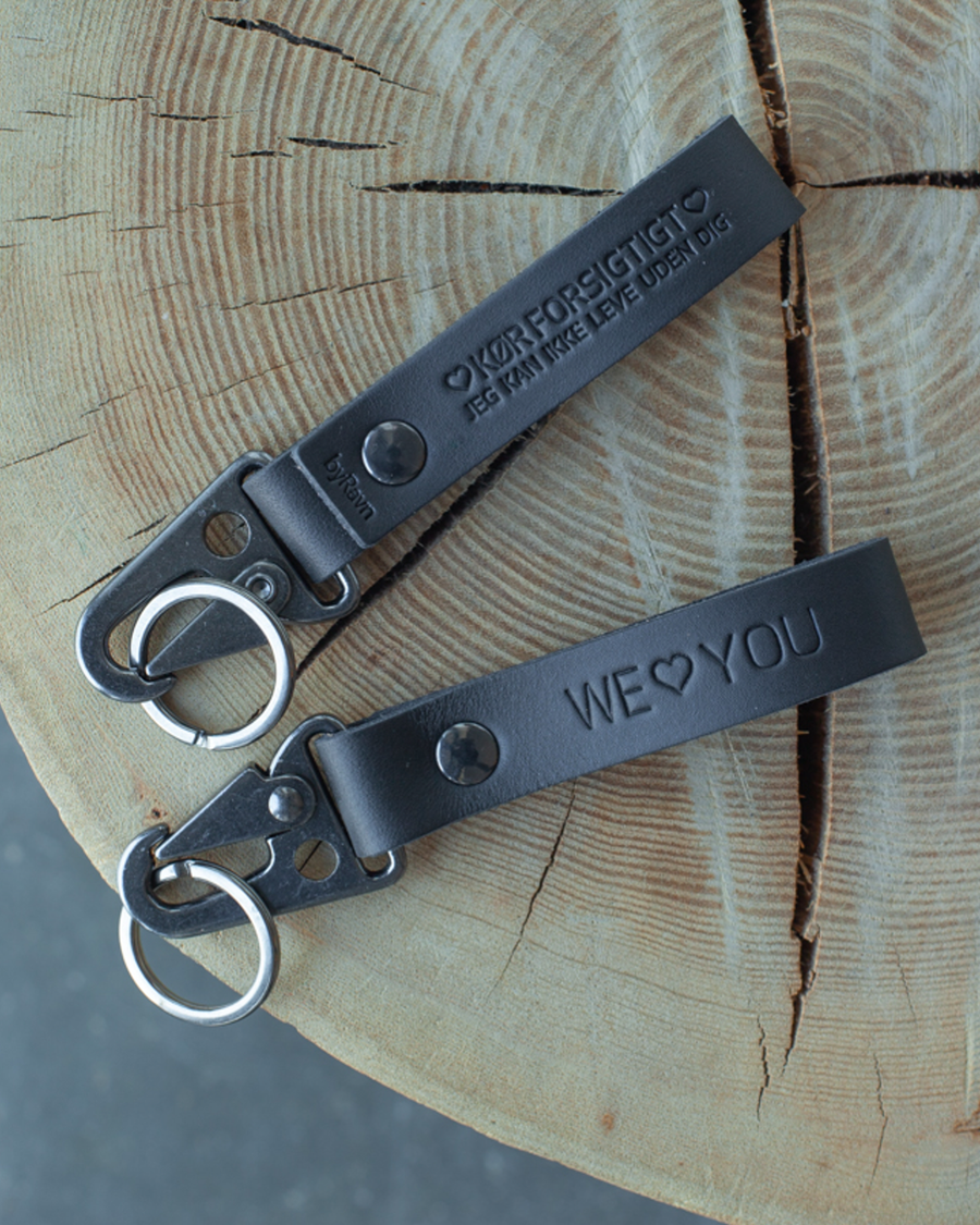 Zwei HOOK Schlüsselanhänger aus schwarzem Leder auf Baumstumpf mit den Gravuren "We ♡ you" und "Kør forsigtigt – jeg kan ikke leve uden dig"
