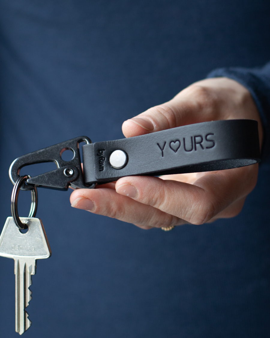 HOOK Schlüsselanhänger aus schwarzem Leder mit der Gravur "Y♡urs" und Karabiner aus schwarzem Stahl, in der Hand gehalten mit Schlüssel