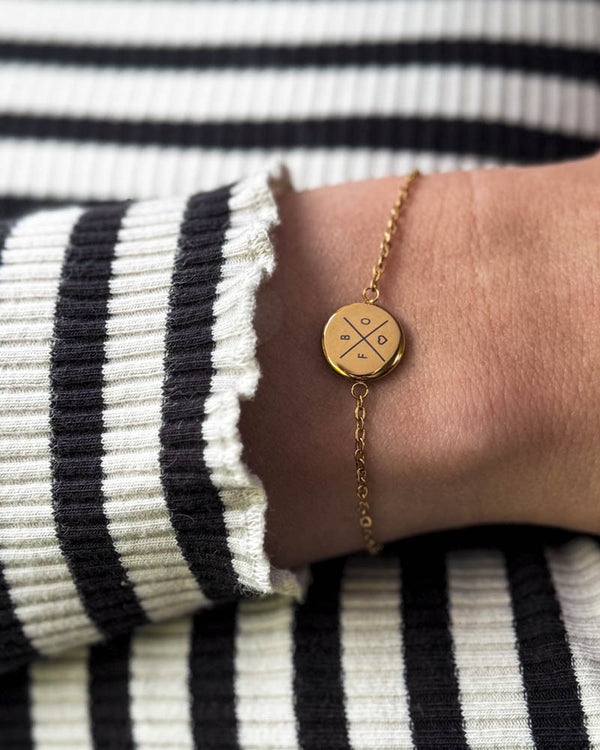 IBI X Armband aus vergoldetem Edelstahl mit Kreuz, Buchstaben und Herz graviert, am Handgelenk zu gestreifter Bluse getragen