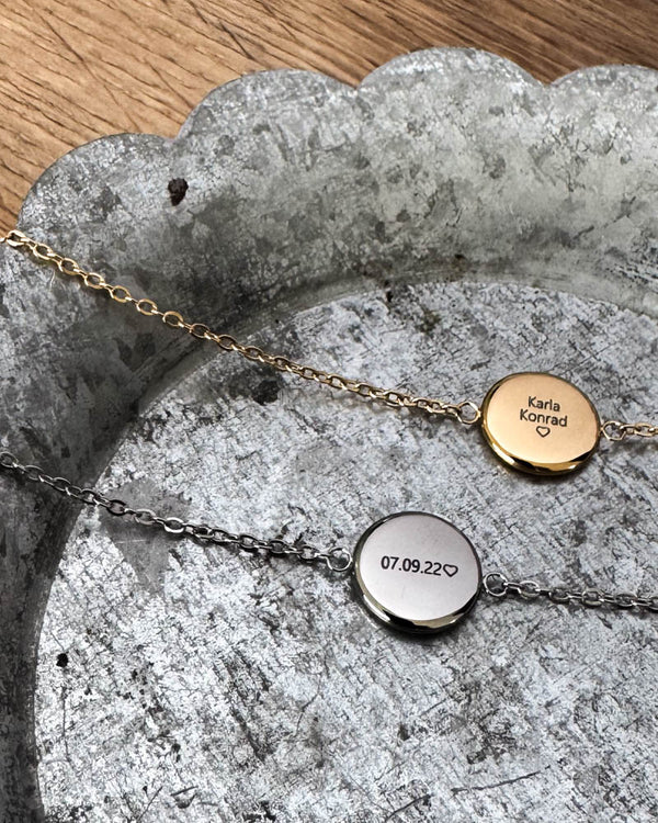 IBI QUOTE Armband aus vergoldetem Edelstahl mit "Karla Konrad" ♡ graviert, neben silberfarbener Variante mit "07.09.22" ♡, auf Zinkschale