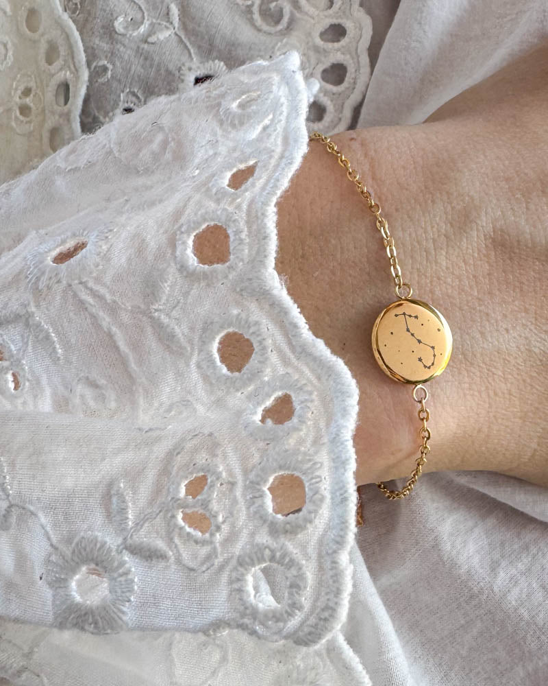 IBI STAR Armband aus vergoldetem Edelstahl mit Sternbild auf runder Platte graviert, am Handgelenk zu weißer Spitzenbluse getragen