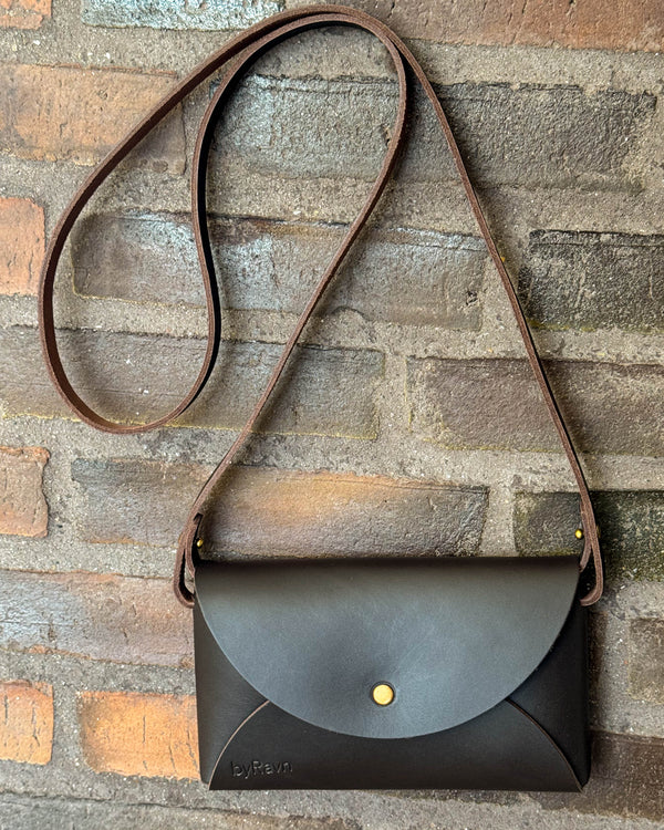 INA Schultertasche aus dunkelbraunem Leder mit langem Riemen, an roter Backsteinwand hängend