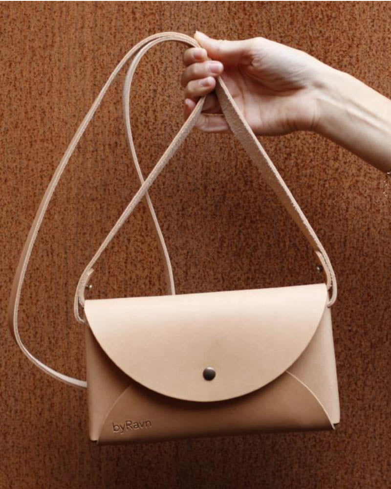 INA Schultertasche aus naturfarbenem Leder, am Riemen hochgehalten vor rostroter Wand
