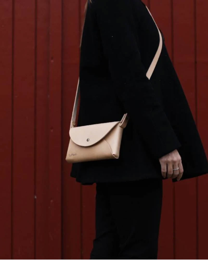 INA Schultertasche aus naturfarbenem Leder, als Crossbody getragen von Frau in Schwarz vor roter Holzverkleidung