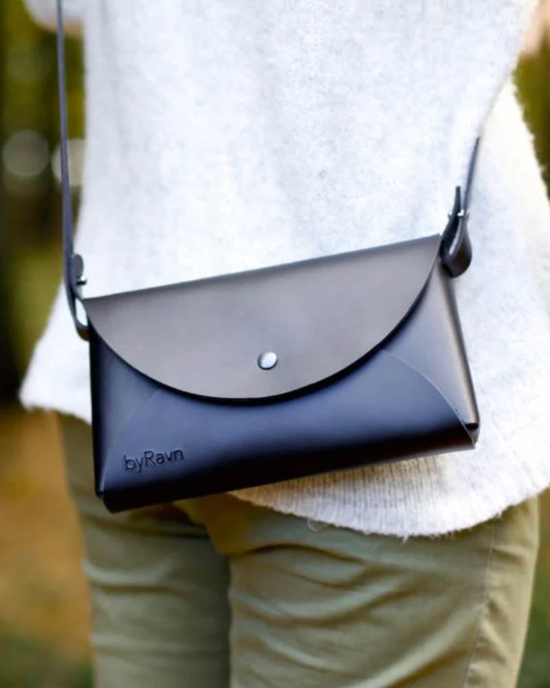 INA Schultertasche aus schwarzem Leder, über der Schulter getragen zu weißem Strickpullover und hellgrüner Hose