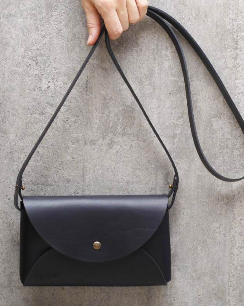 INA Schultertasche aus schwarzem Leder, am Riemen hochgehalten vor grauer Betonwand, mit goldfarbenem Druckknopf