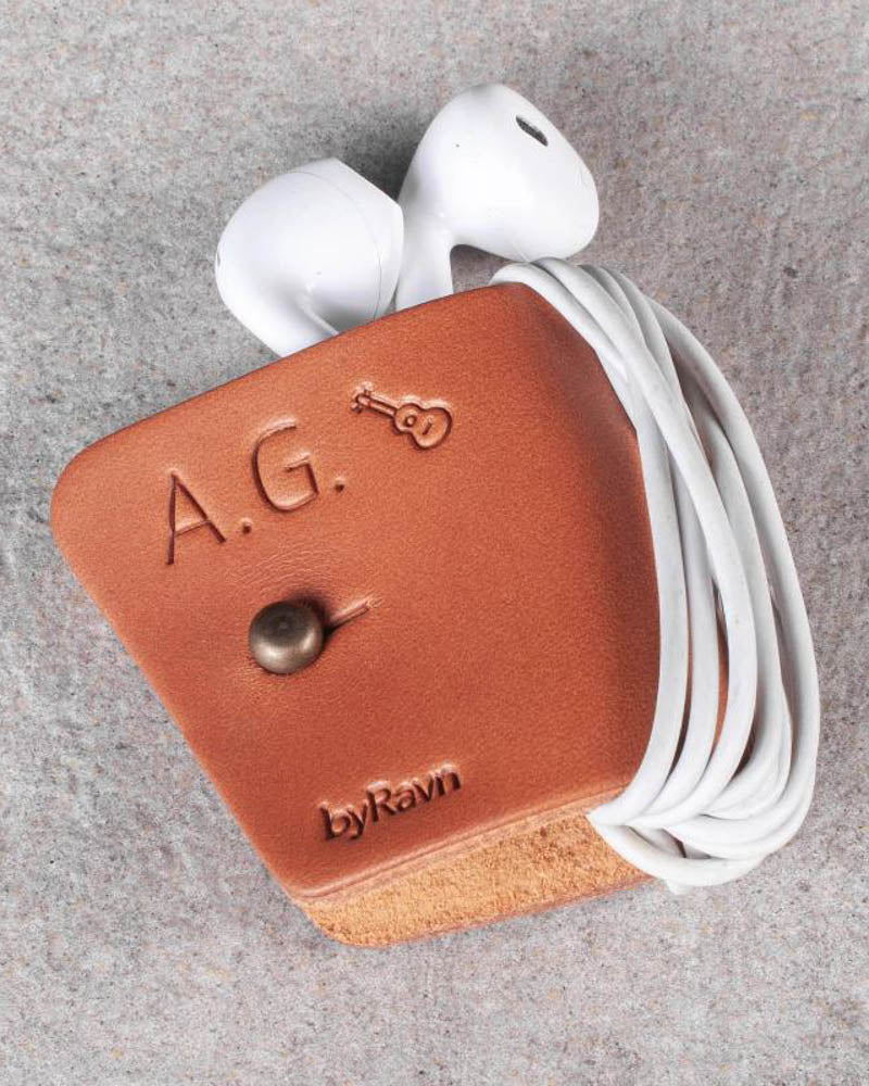 KAX Kopfhörerhalter aus Cognac-Leder mit gravierten Initialen "A.G." und Gitarre, mit Earpods auf grauem Stein