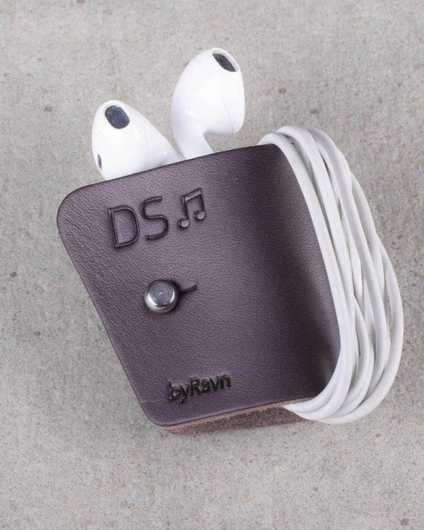 KAX Kopfhörer-Clip aus dunkelbraunem Leder mit umwickelten weißen Earpods und der Gravur "DS" mit Note, auf Betonhintergrund
