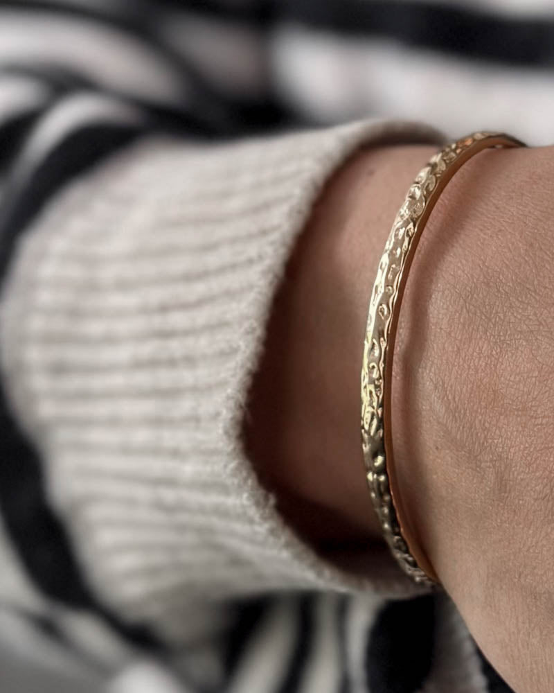 LUNA Armband aus vergoldetem Edelstahl mit gehämmerter Oberfläche am Handgelenk, weißer Strick und gestreifte Bluse