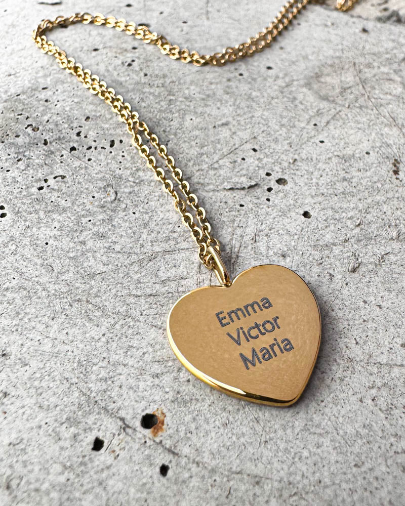 LUV QUOTE Namenskette aus vergoldetem Edelstahl mit den Namen "Emma Victor Maria" auf Herzanhänger graviert, auf Beton liegend