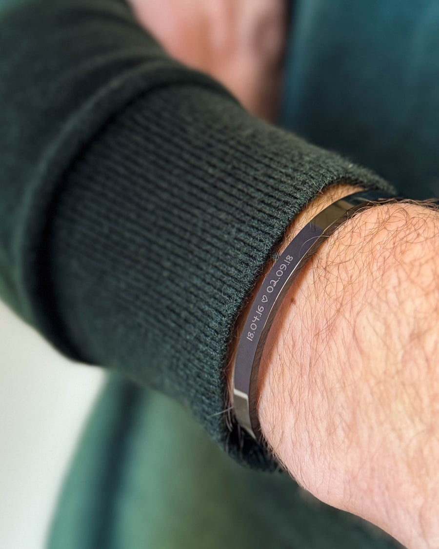 MARS SCRIPT Armband aus schwarzem Edelstahl mit den Daten "18.04.16 ♡ 02.09.18" in Handschrift graviert, zu dunkelgrünem Strick getragen