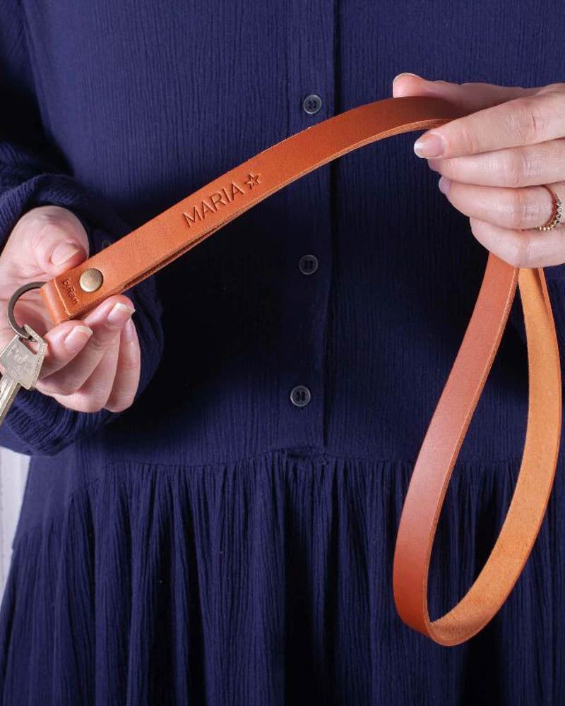 MAX Schlüsselanhänger aus Cognac-Leder, lange Schlaufe mit dem gravierten Namen "Maria" ☆, in den Händen gehalten vor dunkelblauem Kleid