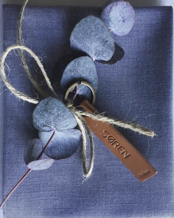 MERRY Tischkarte aus cognacfarbenem Leder mit graviertem Namen "Søren", mit Kordel und Eukalyptus auf blauer Serviette gebunden