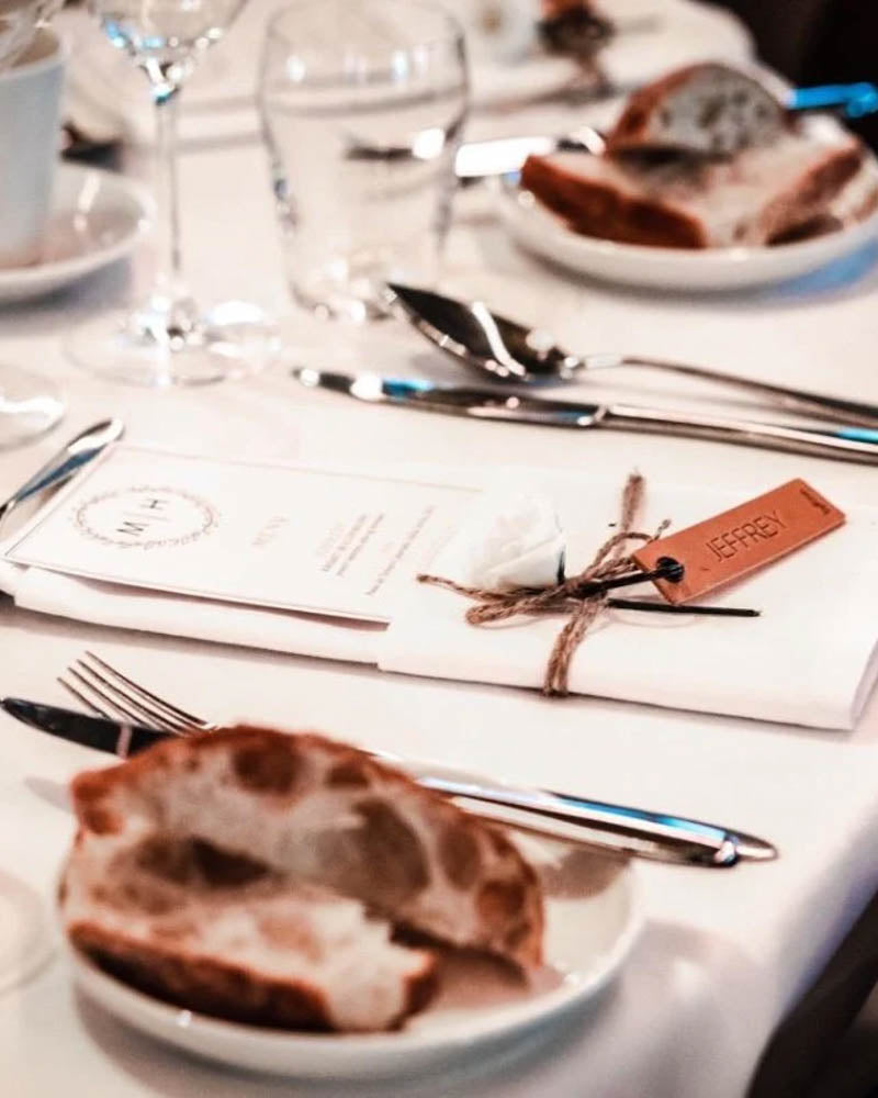 Muster der MERRY Tischkarte aus cognacfarbenem Leder mit graviertem Namen "Jeffrey" neben Menükarte und Brot auf dem Festtisch
