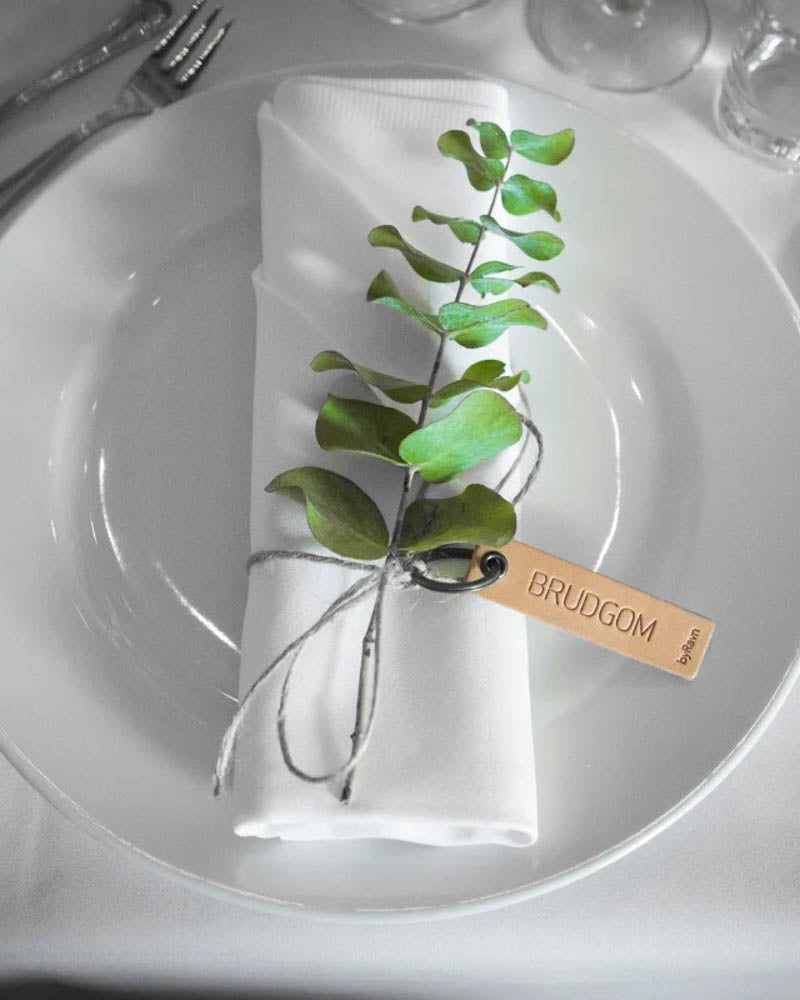MERRY Tischkarte aus naturfarbenem Leder mit der Gravur "Brudgom", um weiße Serviette mit grünem Zweig auf Teller gebunden