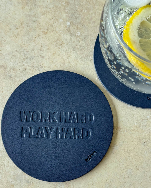 MOKKA Untersetzer aus dunkelblauem Leder mit Prägung "Work hard play hard" auf hellem Tisch neben Glas mit Zitronenscheibe