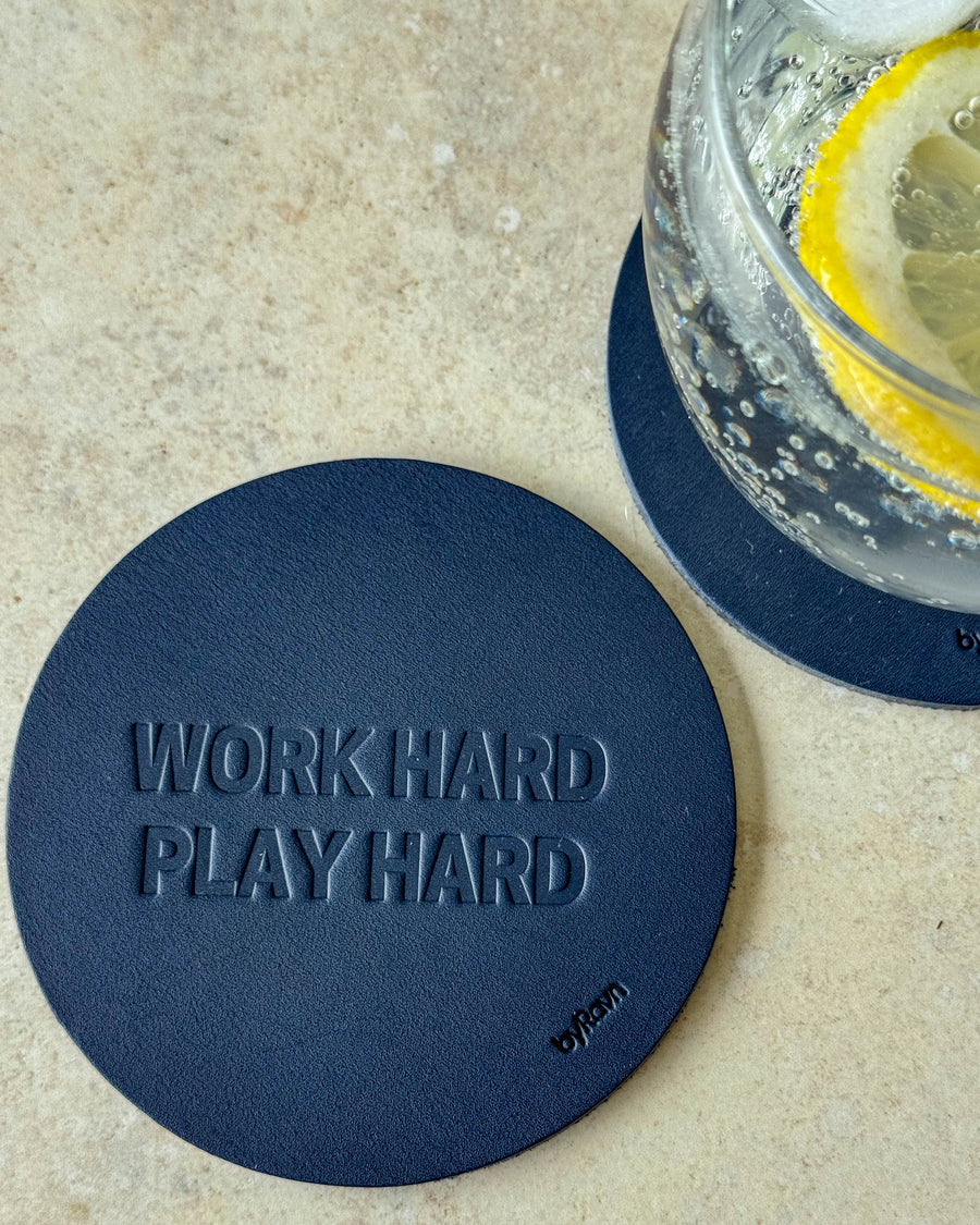 MOKKA Untersetzer aus dunkelblauem Leder mit Prägung "Work hard play hard" auf hellem Tisch neben Glas mit Zitronenscheibe