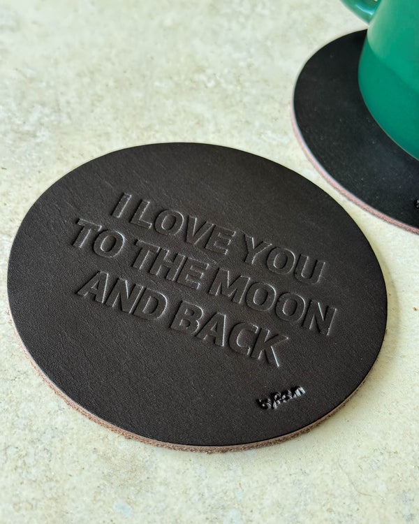 MOKKA Untersetzer aus dunkelbraunem Leder mit Prägung "I love you to the moon and back" auf hellem Tisch neben grüner Tasse