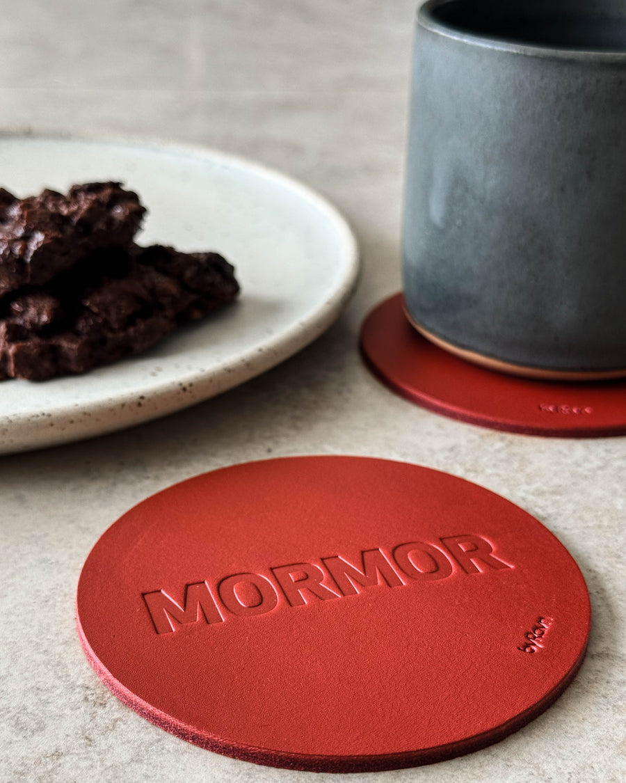 MOKKA Untersetzer aus rotem Leder mit Prägung "Mormor" auf hellem Tisch mit grauer Kaffeetasse und Teller mit Schokoladenkuchen