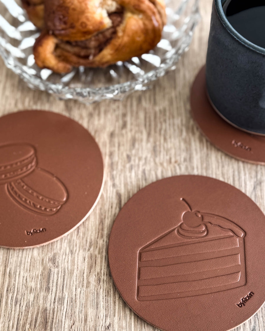 MOKKA Kaffee & Kuchen-Untersetzer aus cognacfarbenem Leder mit geprägter Torte und Macarons auf Holztisch neben Zimtschnecke und schwarzer Tasse
