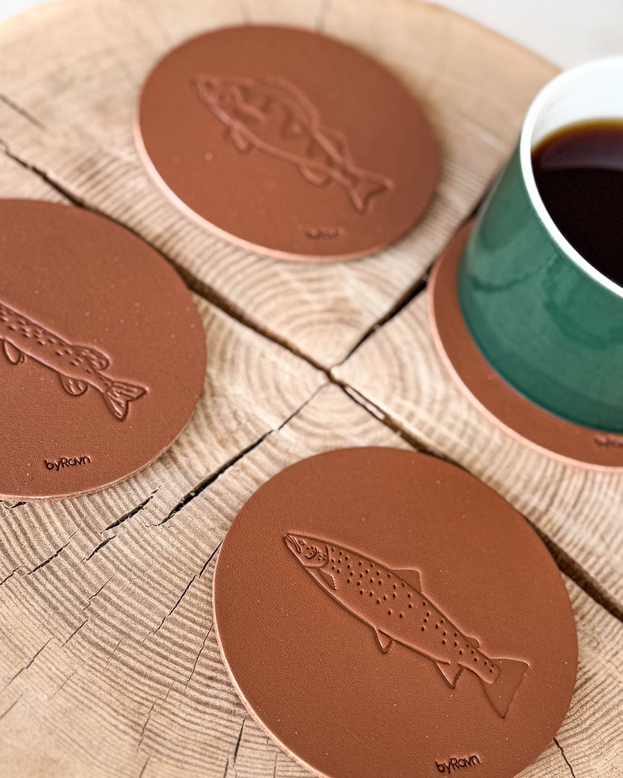 MOKKA Angler-Untersetzer aus cognacfarbenem Leder mit geprägter Meerforelle, Hecht und Barsch auf Holzscheibe neben grüner Kaffeetasse