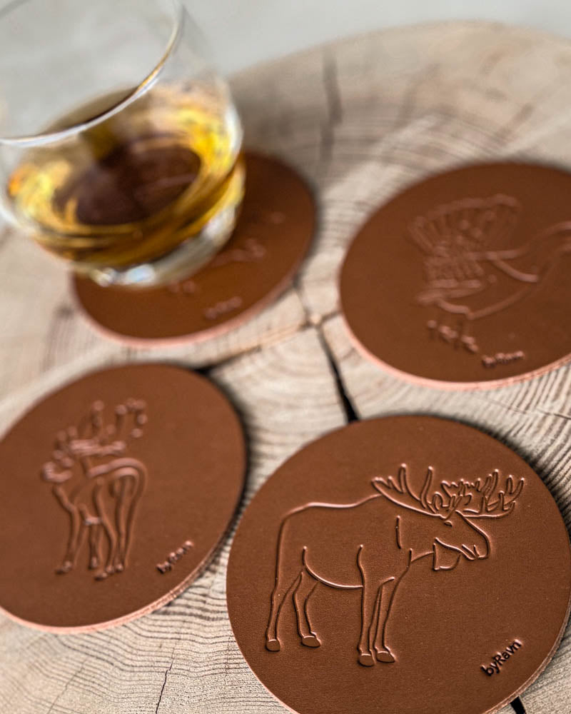 MOKKA Jagd-Untersetzer aus cognacfarbenem Leder mit geprägtem Elch, Damhirsch und Auerhahn auf Holzscheibe neben Glas mit Whisky
