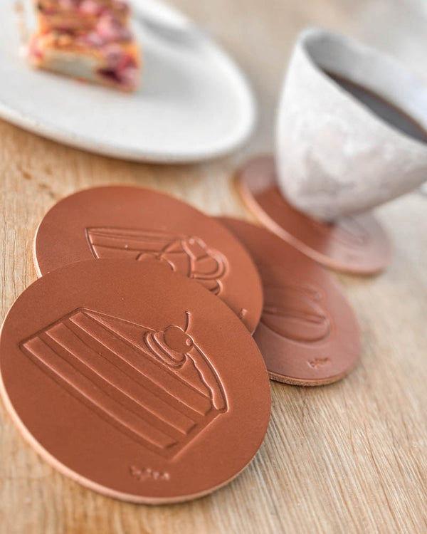 MOKKA Kaffee & Kuchen-Untersetzer aus cognacfarbenem Leder mit geprägter Torte, neben Kaffeetasse und Teller mit Kuchen auf Holztisch