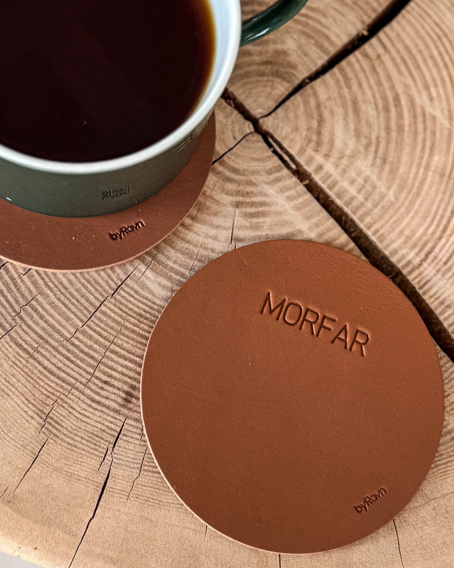 MOKKA runder Untersetzer aus cognacfarbenem Leder mit der Gravur "Morfar" auf Holzscheibe neben grüner Kaffeetasse auf Untersetzer