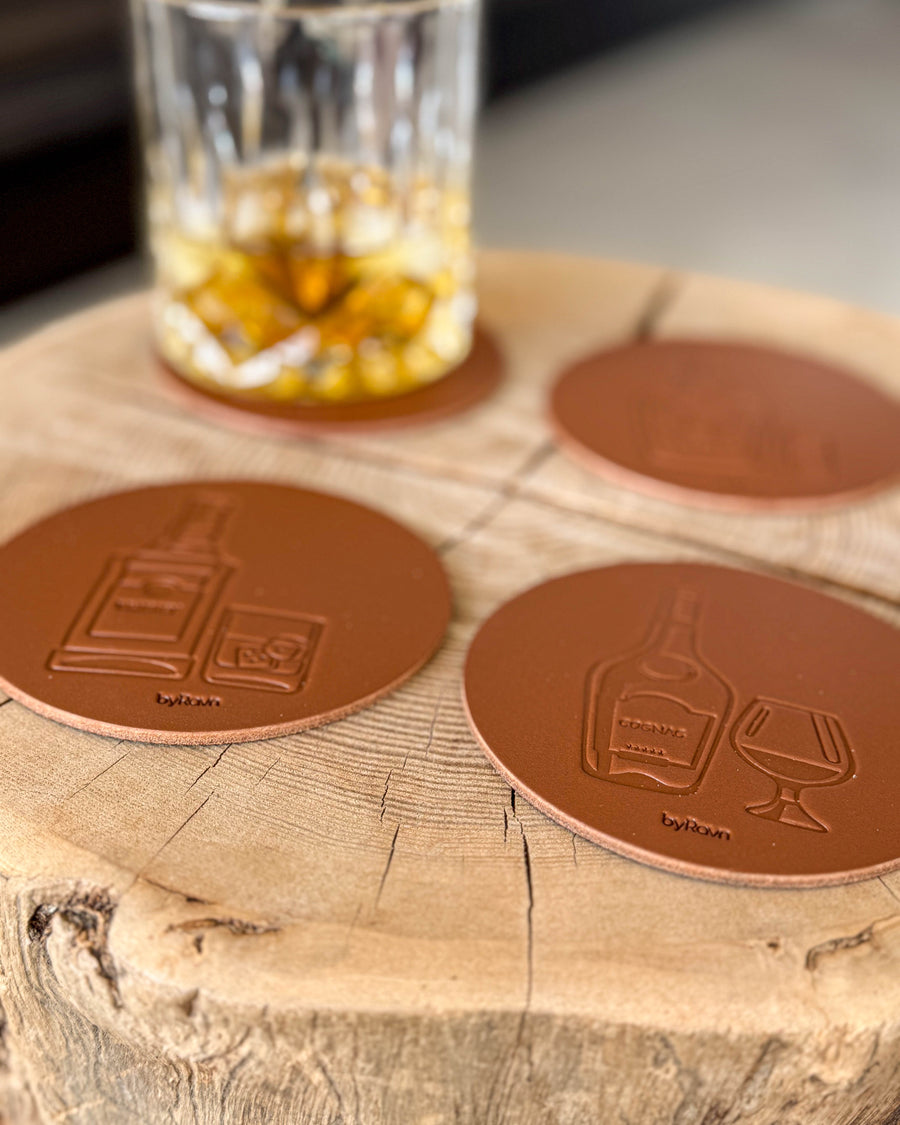MOKKA Spirituosen-Untersetzer aus cognacfarbenem Leder mit Prägung Whisky und Cognac auf Holzscheibe neben Kristallglas mit Whisky und Eis