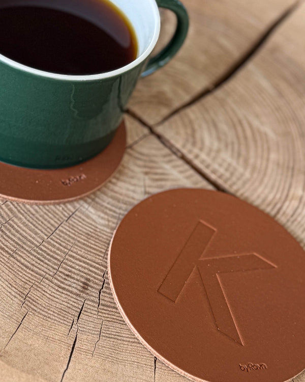 MOKKA Untersetzer aus cognacfarbenem Leder mit geprägtem Buchstaben "K" auf Holzscheibe neben grüner Kaffeetasse auf Untersetzer