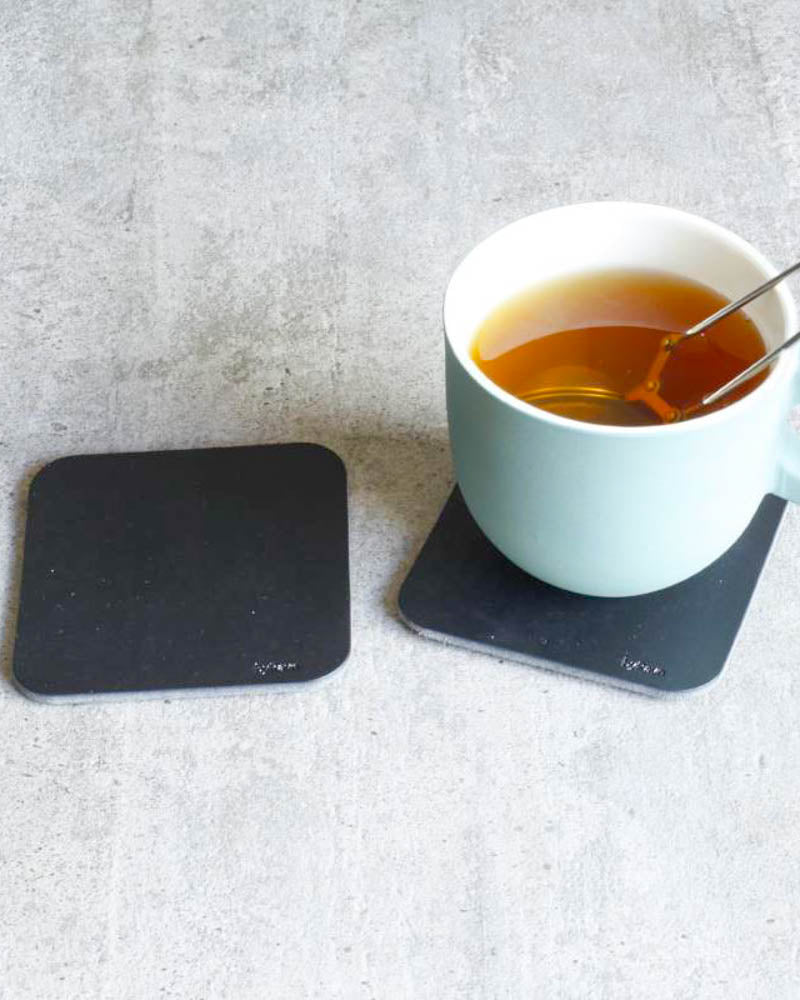 MOKKA quadratische Untersetzer aus schwarzem Leder auf Betontisch, einer unter hellblauer Tasse mit Tee