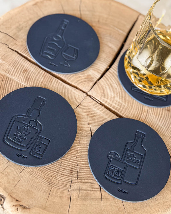 MOKKA Untersetzer aus dunkelblauem Leder mit Spirituosen-Motiven Cognac, Rum und Gin auf Holzscheibe neben Kristallglas mit Whisky und Eis