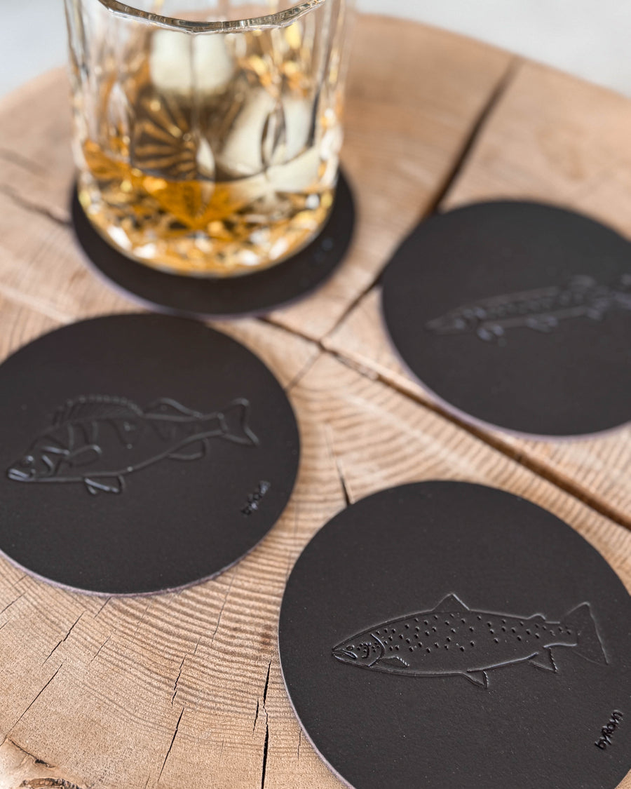 MOKKA Angler-Untersetzer aus dunkelbraunem Leder mit geprägter Meerforelle, Barsch und Hecht auf Holzscheibe neben Whiskyglas