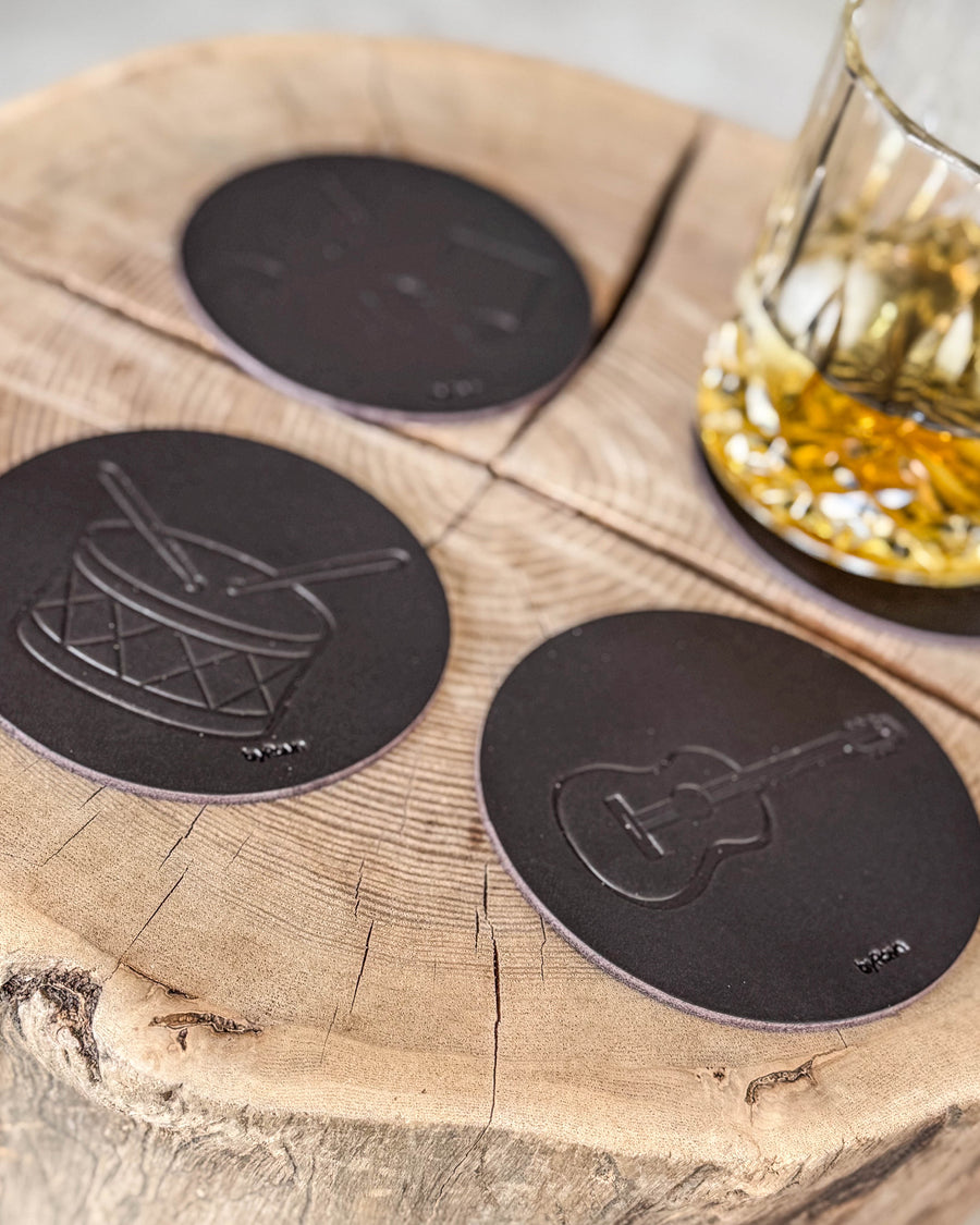 MOKKA Untersetzer aus dunkelbraunem Leder, geprägt mit Trommel und Gitarre, auf Holzscheibe neben Kristallglas mit Whisky