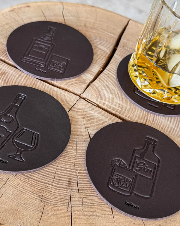 MOKKA Spirituosen-Untersetzer aus dunkelbraunem Leder mit Prägung Gin, Cognac und Whisky auf Holzscheibe neben Glas mit Whisky und Eis