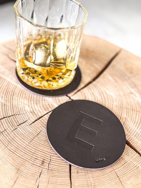MOKKA Untersetzer aus dunkelbraunem Leder mit geprägtem Buchstaben "E" auf Holzscheibe neben Whiskyglas auf Untersetzer
