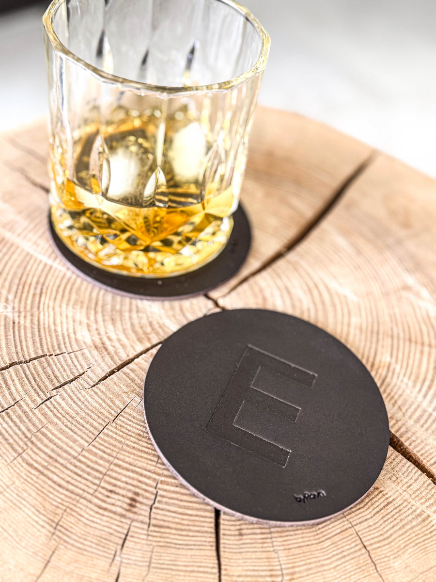 MOKKA Untersetzer aus dunkelbraunem Leder mit geprägtem Buchstaben "E" auf Holzscheibe neben Whiskyglas auf Untersetzer