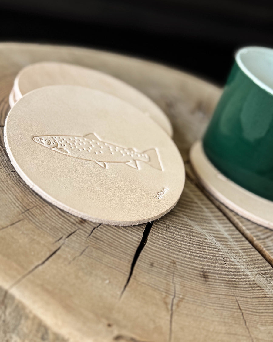 MOKKA Angler-Untersetzer aus naturfarbenem Leder mit geprägter Meerforelle auf Holztisch neben grüner Tasse auf Untersetzer