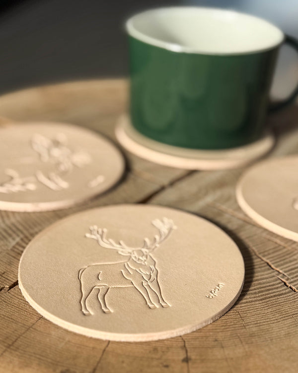 MOKKA Jagd-Untersetzer aus naturfarbenem Leder mit geprägtem Rothirsch auf Holztisch, weitere Untersetzer und grüne Tasse dahinter