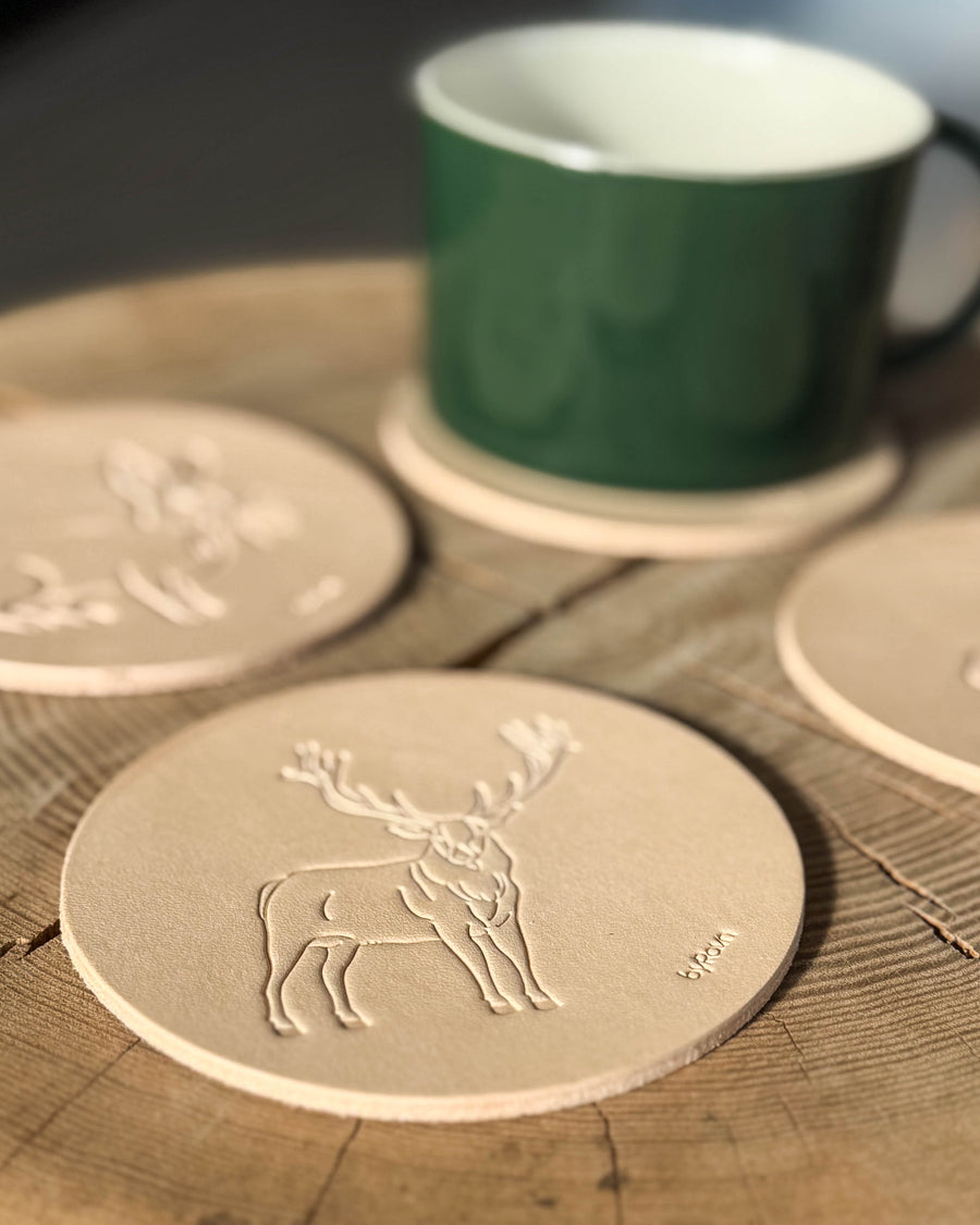 MOKKA Jagd-Untersetzer aus naturfarbenem Leder mit geprägtem Rothirsch auf Holztisch, weitere Untersetzer und grüne Tasse dahinter