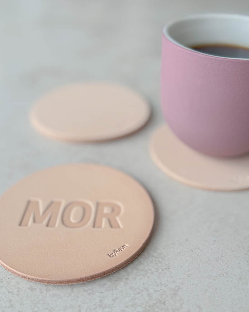 MOKKA Untersetzer aus naturfarbenem Leder mit Prägung "Mor" in Großbuchstaben, rosa Kaffeetasse auf Untersetzer dahinter