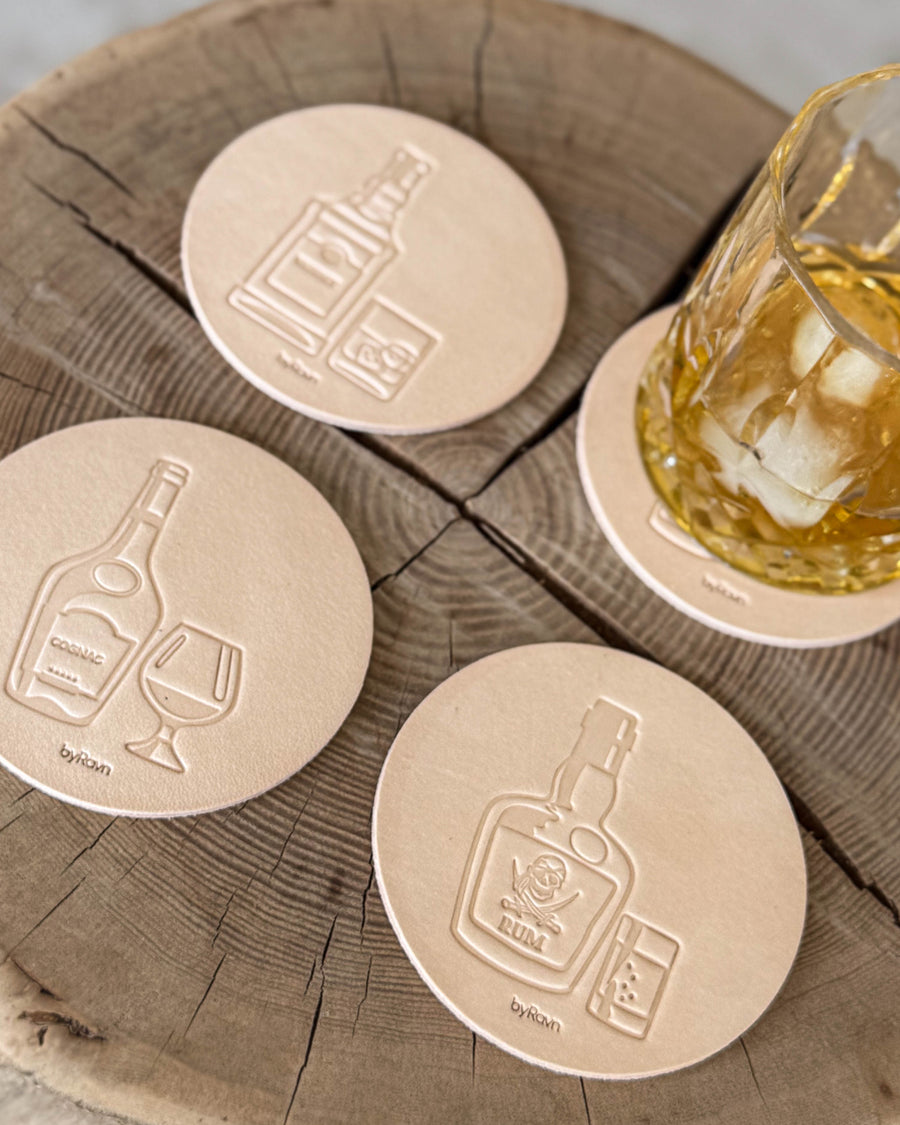 MOKKA Spirituosen-Untersetzer aus naturfarbenem Leder mit Prägung Cognac, Rum und Whisky auf Holzscheibe neben Glas mit Whisky und Eis