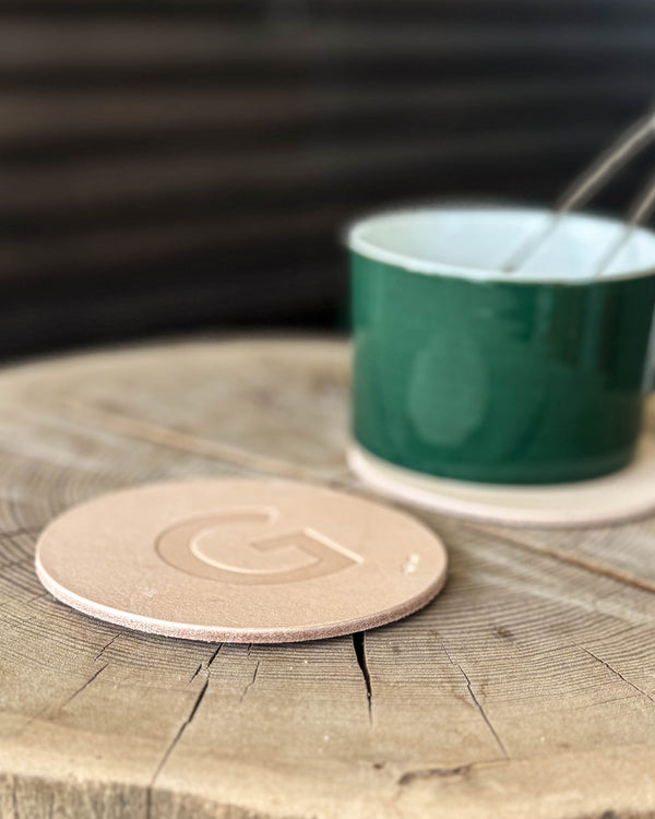 MOKKA Untersetzer aus naturfarbenem Leder mit geprägtem Buchstaben "G" auf Holzscheibe vor grüner Tasse mit Löffel