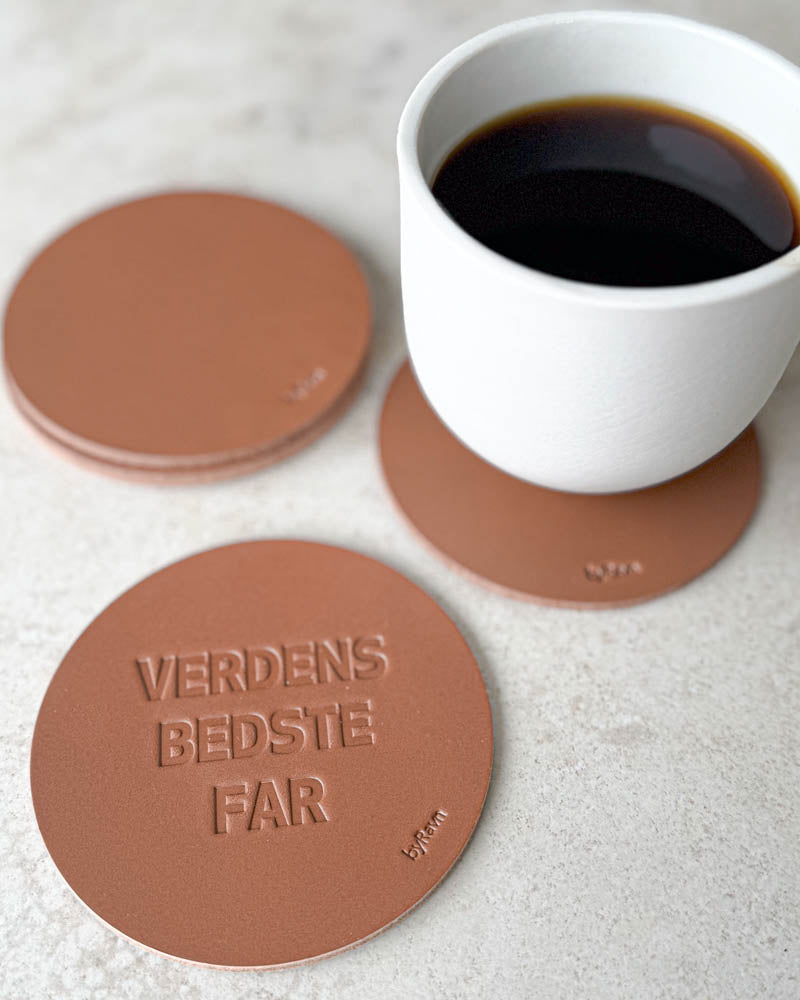 MOKKA Untersetzer aus cognacfarbenem Leder mit Prägung "Verdens bedste far" auf hellem Tisch mit weißer Kaffeetasse