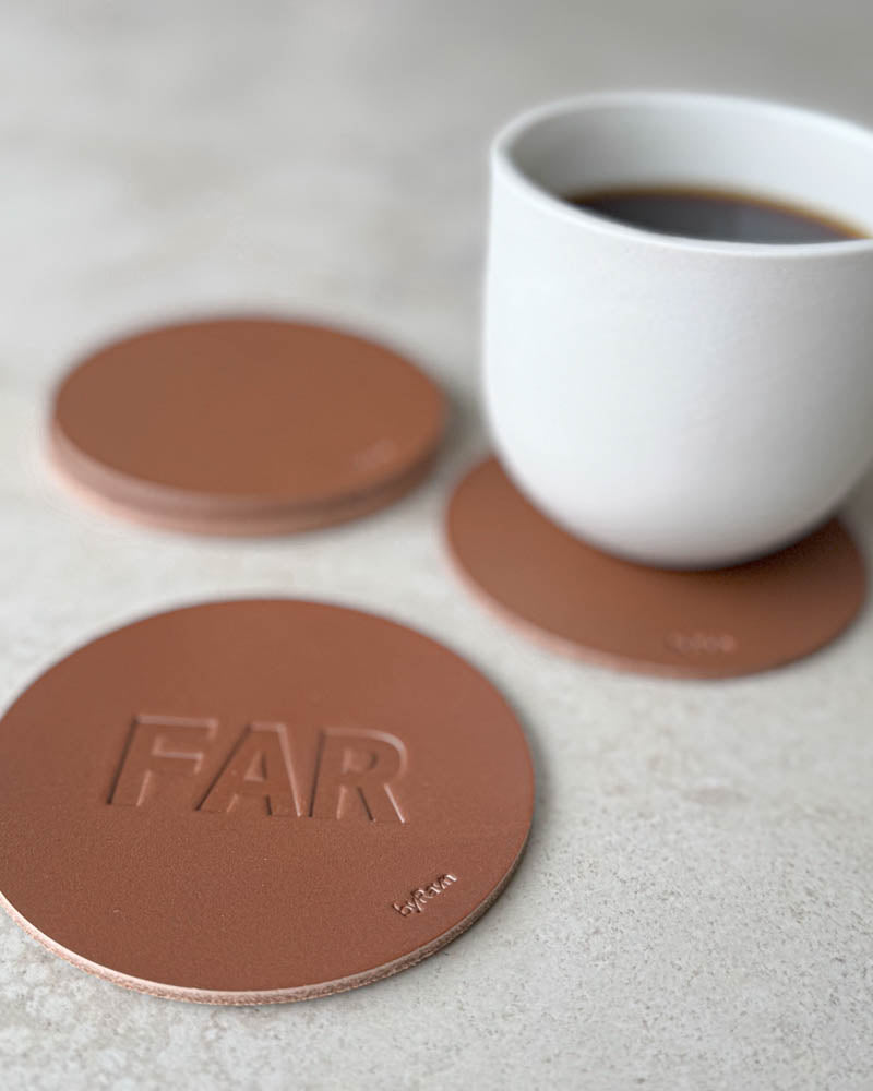 MOKKA Untersetzer aus cognacfarbenem Leder mit Prägung "Far" in Großbuchstaben, weitere Untersetzer und weiße Kaffeetasse dahinter