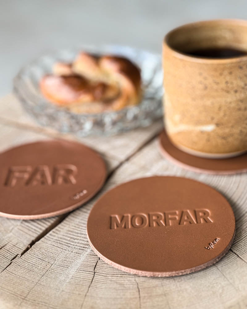 MOKKA Untersetzer aus cognacfarbenem Leder mit Prägung "Far" und "Morfar" auf Holzscheibe mit Kaffeetasse und Zimtschnecke auf Glasteller