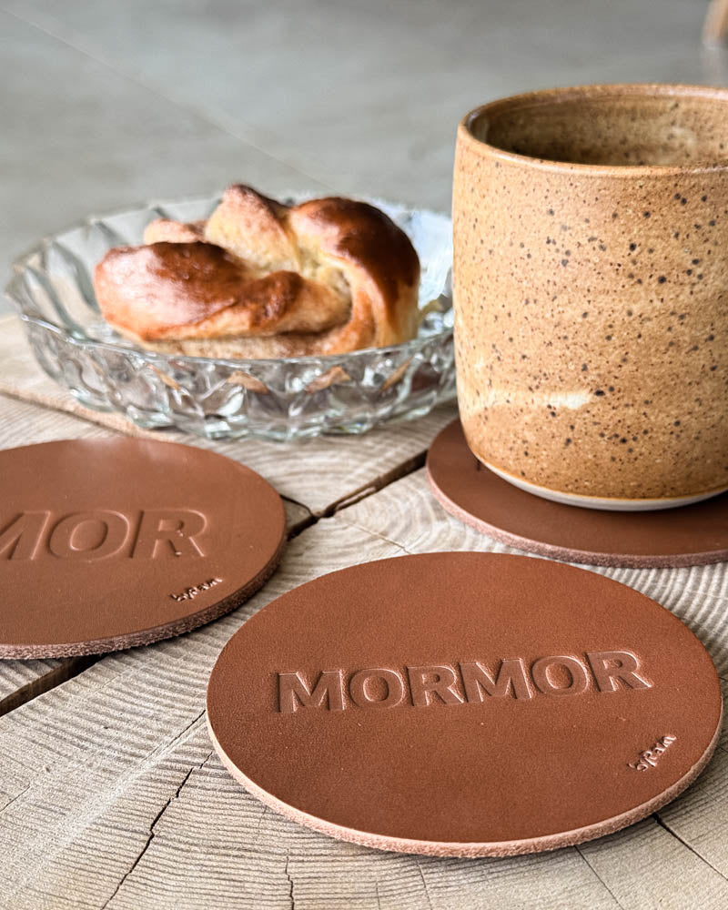 MOKKA Untersetzer aus cognacfarbenem Leder mit Prägung "Mor" und "Mormor" auf Holztisch mit Kaffeetasse und Zimtschnecke in Glasschale